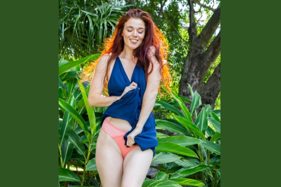 A smiling woman with long, flowing red hair wearing coral lace underwear and a navy wrap top, standing amidst lush greenery.