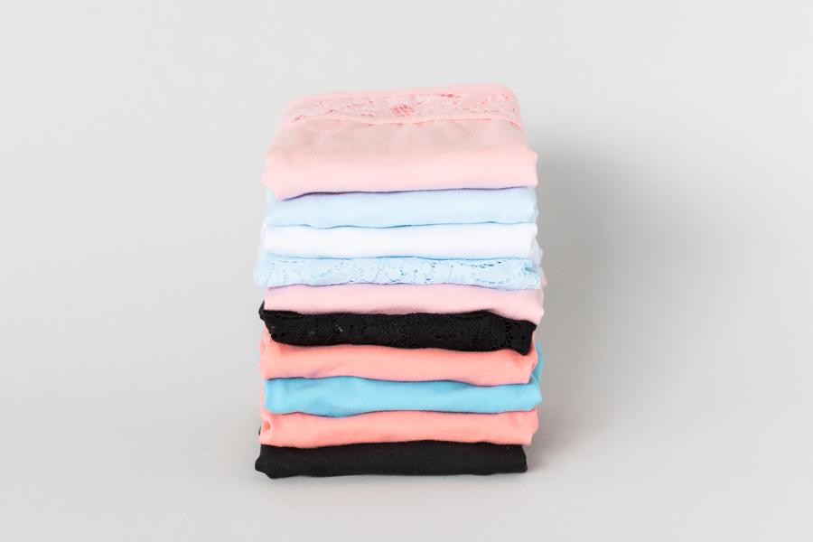 A neatly stacked pile of women's underwear in pastel shades of pink, blue, and peach with lace detailing, against a white background.